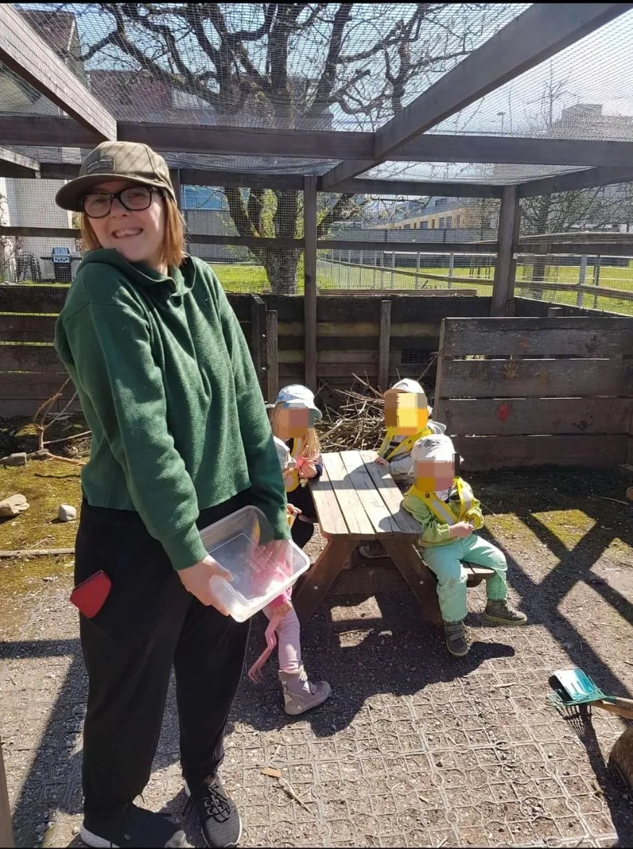 Nadia mit einer Dose vor einer Vogelvoliere. Neben ihr sitzen Kinder an einem kleinen Tisch; ihre Gesichter sind verpixelt.