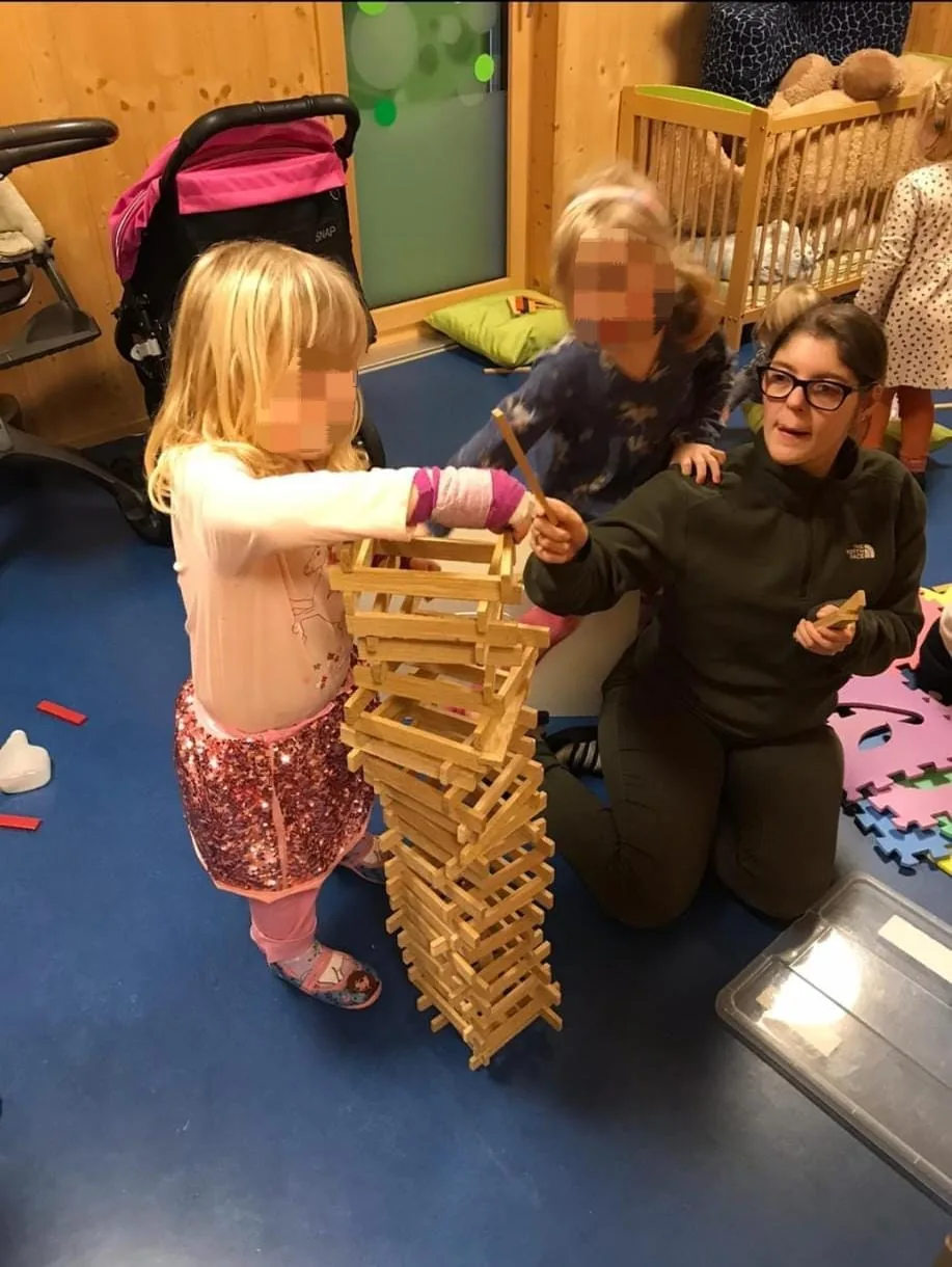 Nadia baut mit Kindern einen Turm aus Holzklötzen. Sichtbare Kindergesichter sind unkenntlich gemacht.