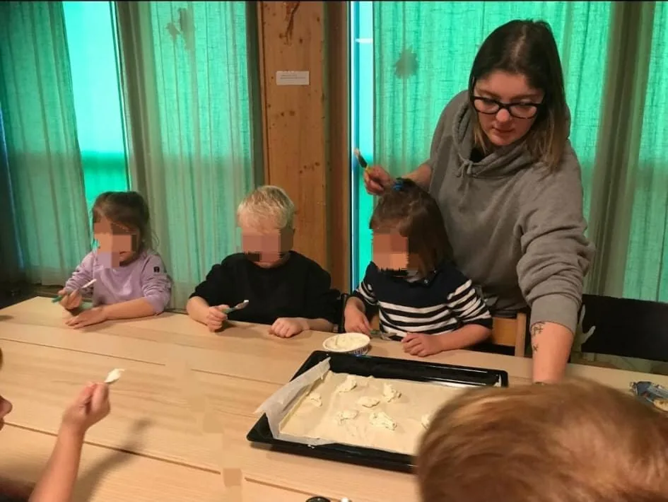 Nadia steht mit einem Löffel neben Kindern an einem Tisch mit Backblech. Sichtbare Kindergesichter sind verpixelt.